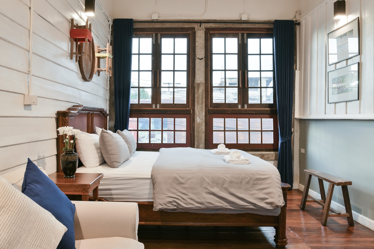 A serene room features a spacious bed with neutral bedding, flanked by large windows that allow ample natural light. Blue curtains frame the windows, and a small seating area is visible with a comfortable armchair. A simple wooden bench and decor elements add to the charm.