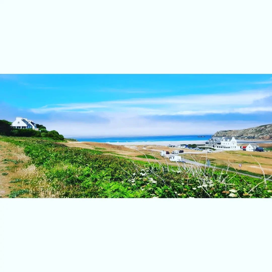 A scenic coastal view showcases rolling hills and sandy shores under a clear blue sky. White houses dot the landscape, with the ocean visible in the background, enhancing the tranquility of the area. Greenery lines the foreground, adding to the natural beauty of the scene.