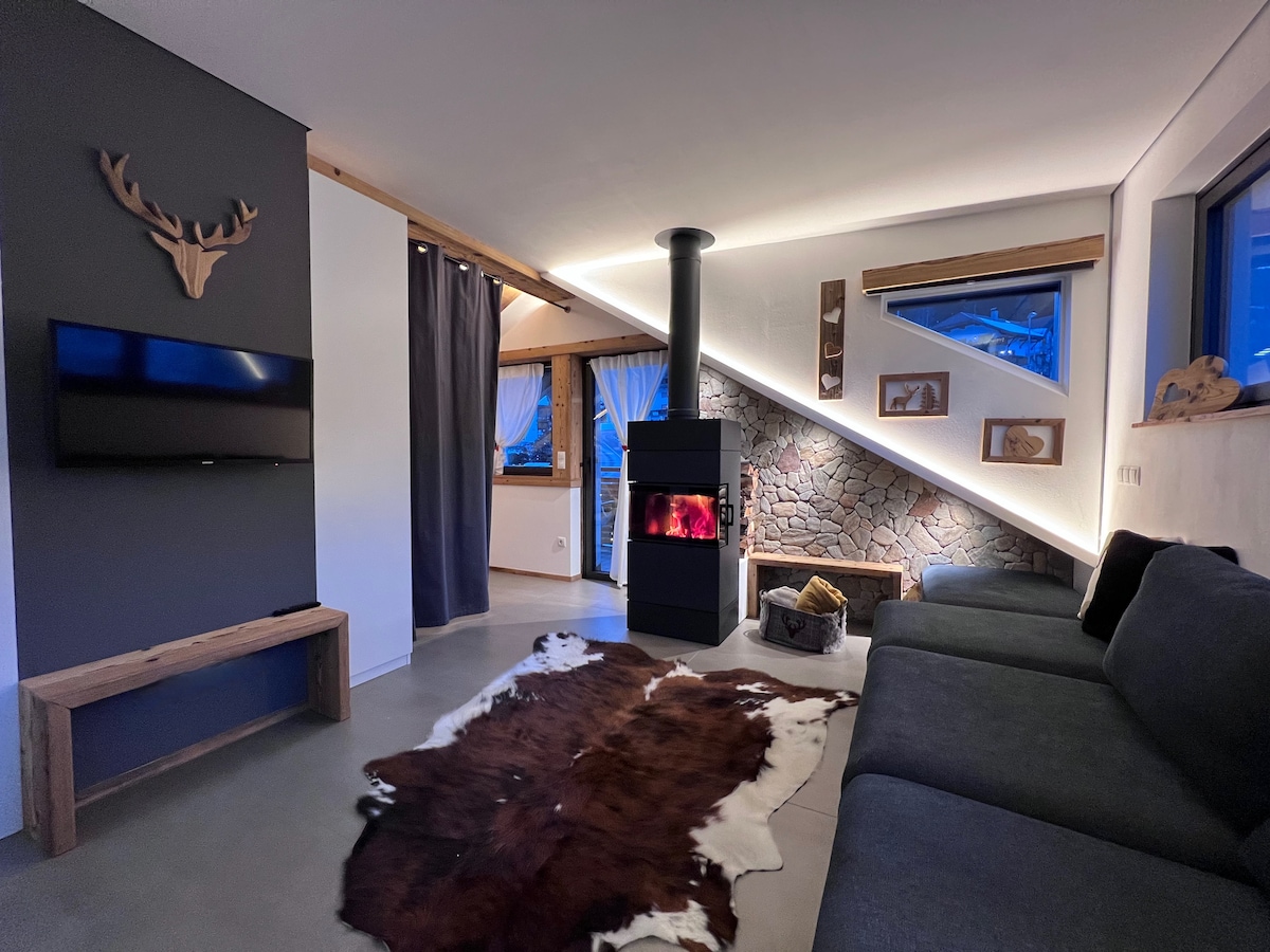 A modern living area is highlighted by a sleek wood-burning stove situated against a stone wall. A large dark sofa offers comfortable seating, accompanied by a decorative cowhide rug. A mounted television and art pieces accent the walls, with natural light filtering through the triangular window.
