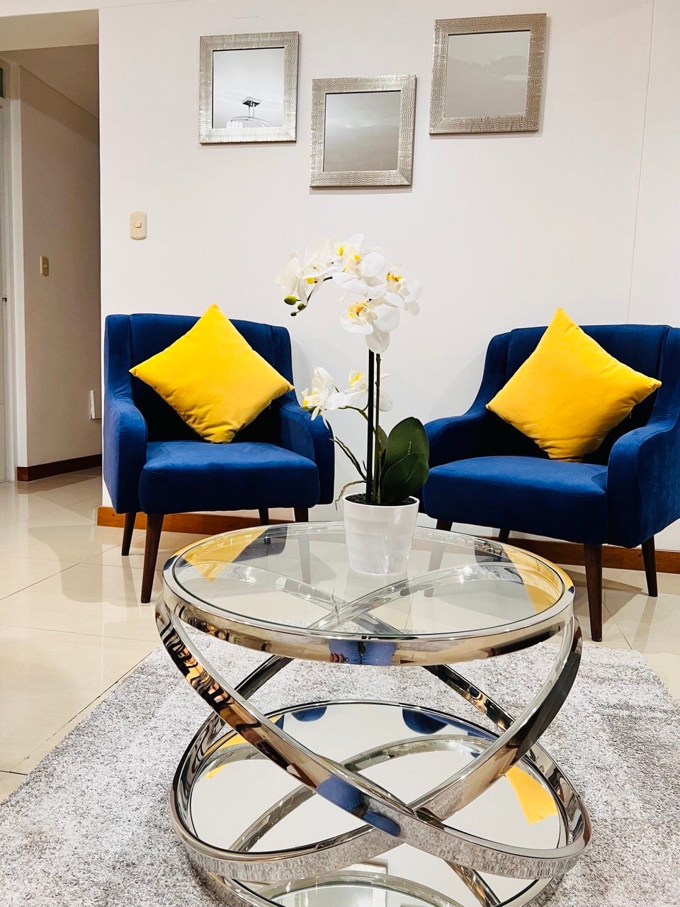 A seating area features two blue armchairs with yellow cushions, positioned around a round, reflective coffee table. A white orchid in a simple pot rests on the table, contributing to the elegant decor. Three framed mirrors adorn the wall above the seating.