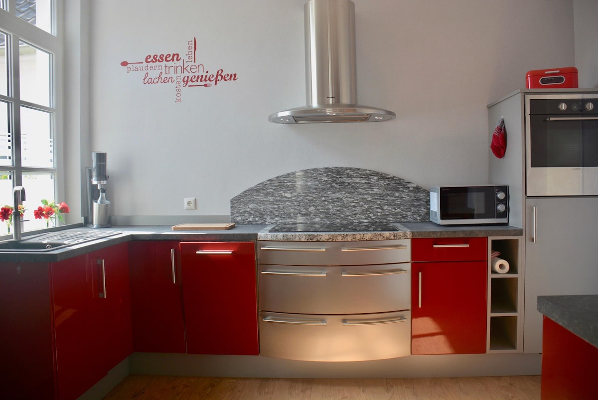 The modern kitchen features red and gray cabinetry, complemented by a stylish granite countertop. Essential appliances, including a microwave and oven, are built-in for convenience. Natural light floods the space through the large window, enhancing the bright and functional design.