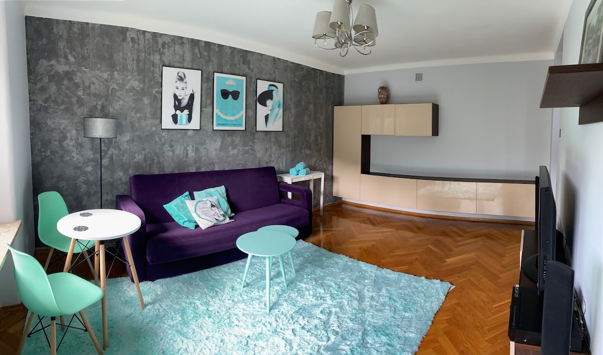 A modern living area features a purple sofa against a textured grey wall. A light blue rug covers the floor, complemented by a white coffee table and matching side tables. Artwork is displayed on the wall, and a television stand is positioned nearby.