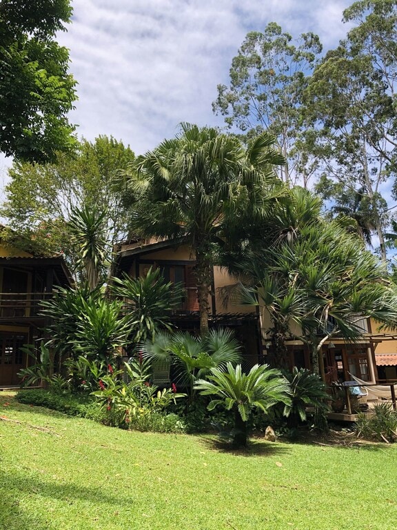 The exterior of the property is surrounded by lush greenery and tropical plants, with tall trees providing shade. The multi-story structure blends naturally into the environment, showcasing wooden accents that enhance its charm. A spacious lawn area invites outdoor relaxation.