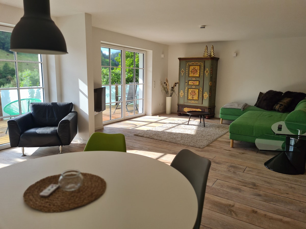 A bright open living area is featured, showcasing large glass doors that invite natural light and views of the greenery outside. A mix of seating options, including a green sectional sofa and black armchair, complements the dining table set for four. Traditional decor adds character to the space.