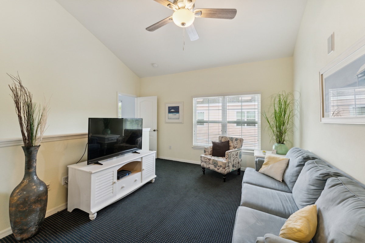 A well-lit living area showcases a ceiling fan and light-colored walls. A gray sofa and patterned armchair provide seating options, while a white media console holds a television. Large windows allow natural light to fill the space, and decorative plants add a touch of greenery.