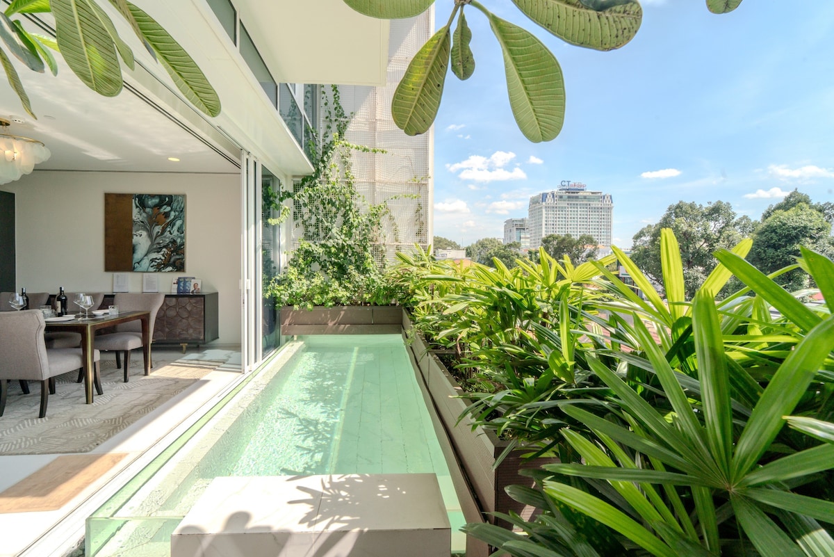 An outdoor balcony is visible, surrounded by lush greenery. A small private pool runs along one side, reflecting the clear blue sky. The dining area indoors can be glimpsed through expansive glass doors, enhancing the connection between indoor and outdoor spaces.