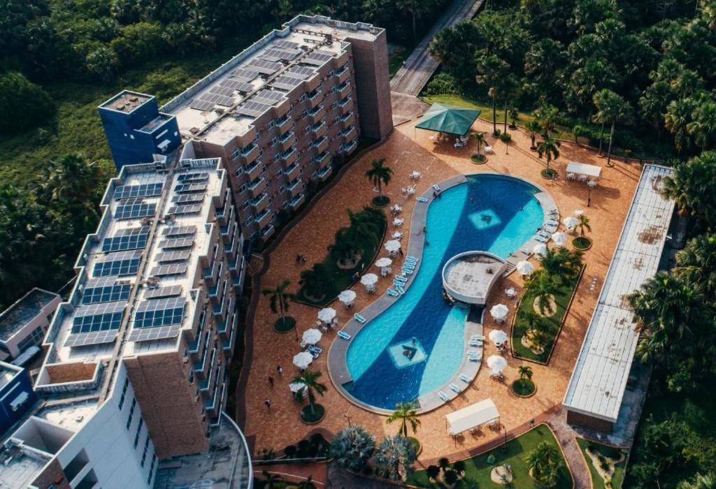 An aerial view captures the resort's expansive layout, featuring a large swimming pool with curved edges and surrounded by lounge chairs. Lush greenery borders the property, and the building's modern architecture is highlighted by solar panels on the roof.