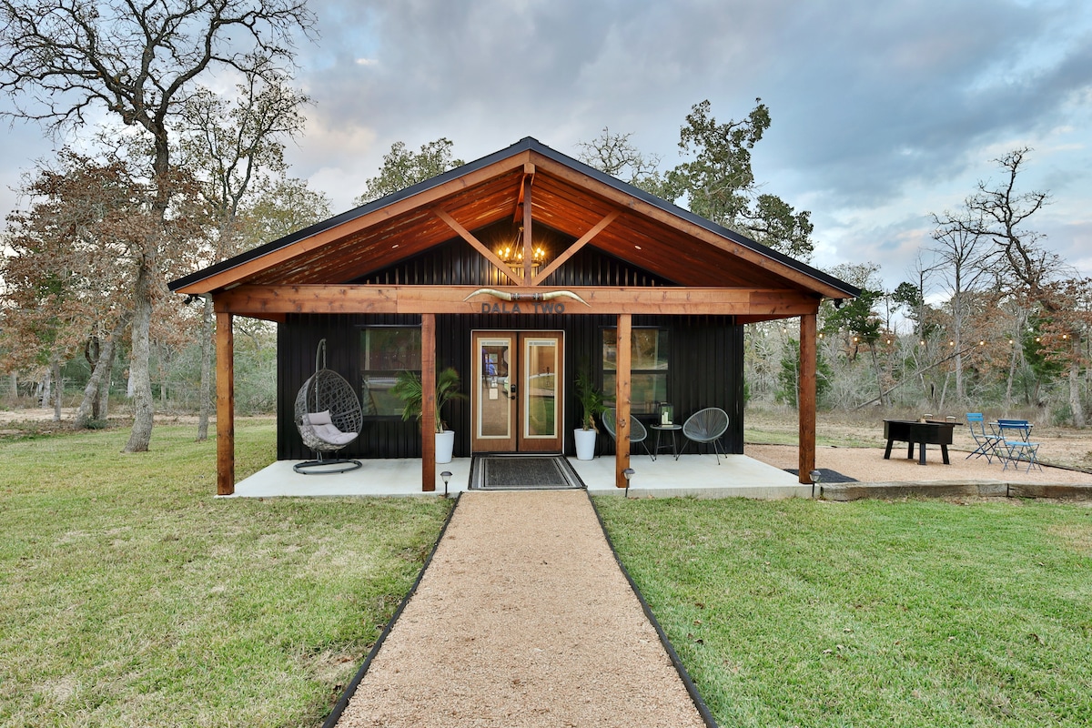 The entrance of a cozy cottage features a large overhang supported by wooden beams. Two hanging chairs are positioned on the porch, surrounded by potted plants. A smooth gravel path leads to the front door, with a grassy area extending to the sides.