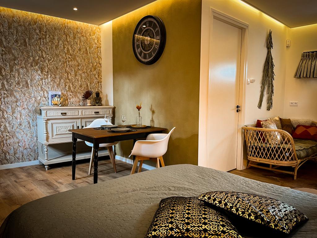 A thoughtfully arranged interior features a spacious bed covered with decorative cushions, a stylish wooden desk beside a vintage sideboard, and a cozy seating area with a rattan chair. The warm-toned walls are complemented by tasteful wall art and subtle lighting.