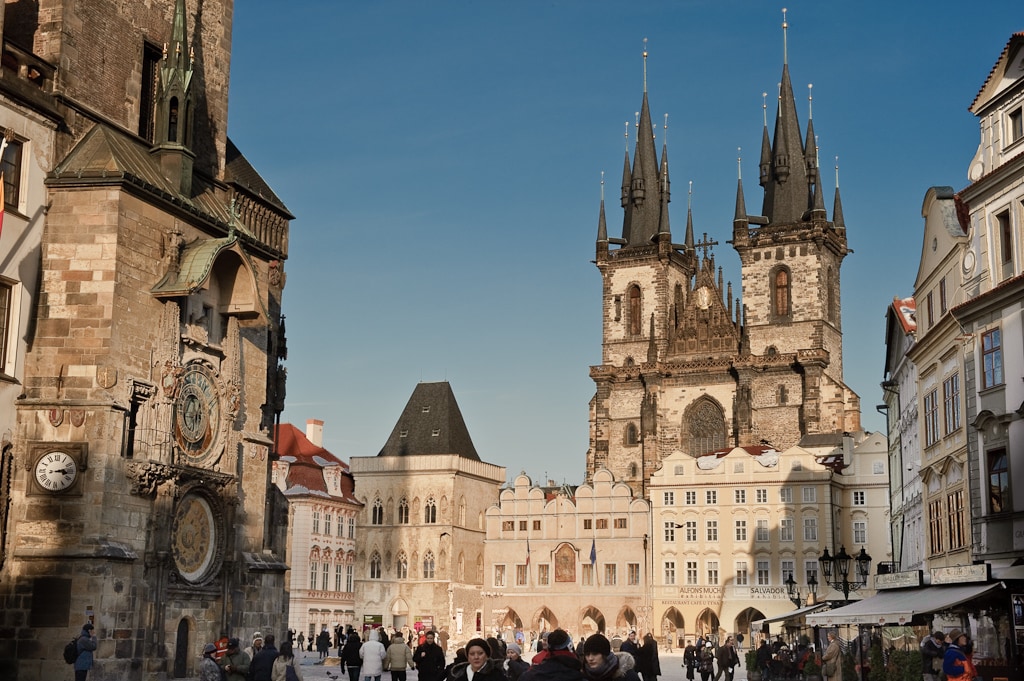 The image captures a historic square featuring the Týn Church with its distinctive spires against a clear blue sky. Surrounding buildings showcase ornate architecture, hinting at the rich history of the area, while pedestrians add a lively atmosphere to the scene.