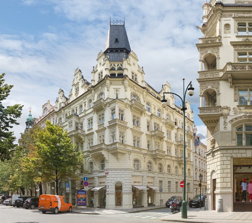 The exterior of a historic building is showcased, featuring ornate architectural details and a central tower. Surrounding trees provide a hint of greenery, contributing to a serene environment. Nearby streets are visible, with signage and a parked vehicle adding to the urban scene.