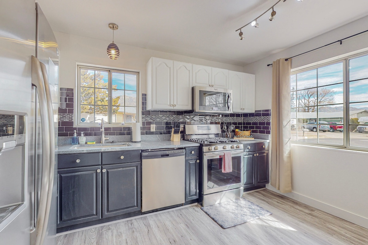 A modern kitchen is presented with dark cabinetry and stainless steel appliances, including a gas range and dishwasher. Natural light enters through a large window, highlighting the sleek countertop and decorative backsplash. A small rug is placed in front of the sink, adding warmth to the space.