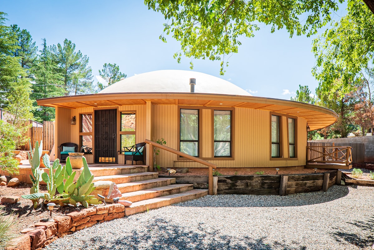The unique dome-shaped house is surrounded by lush greenery, featuring a spacious front porch with steps leading to the entrance. Large windows are visible, allowing natural light to fill the interior. A simple desert landscape with rock formations and cacti can be seen in the foreground.