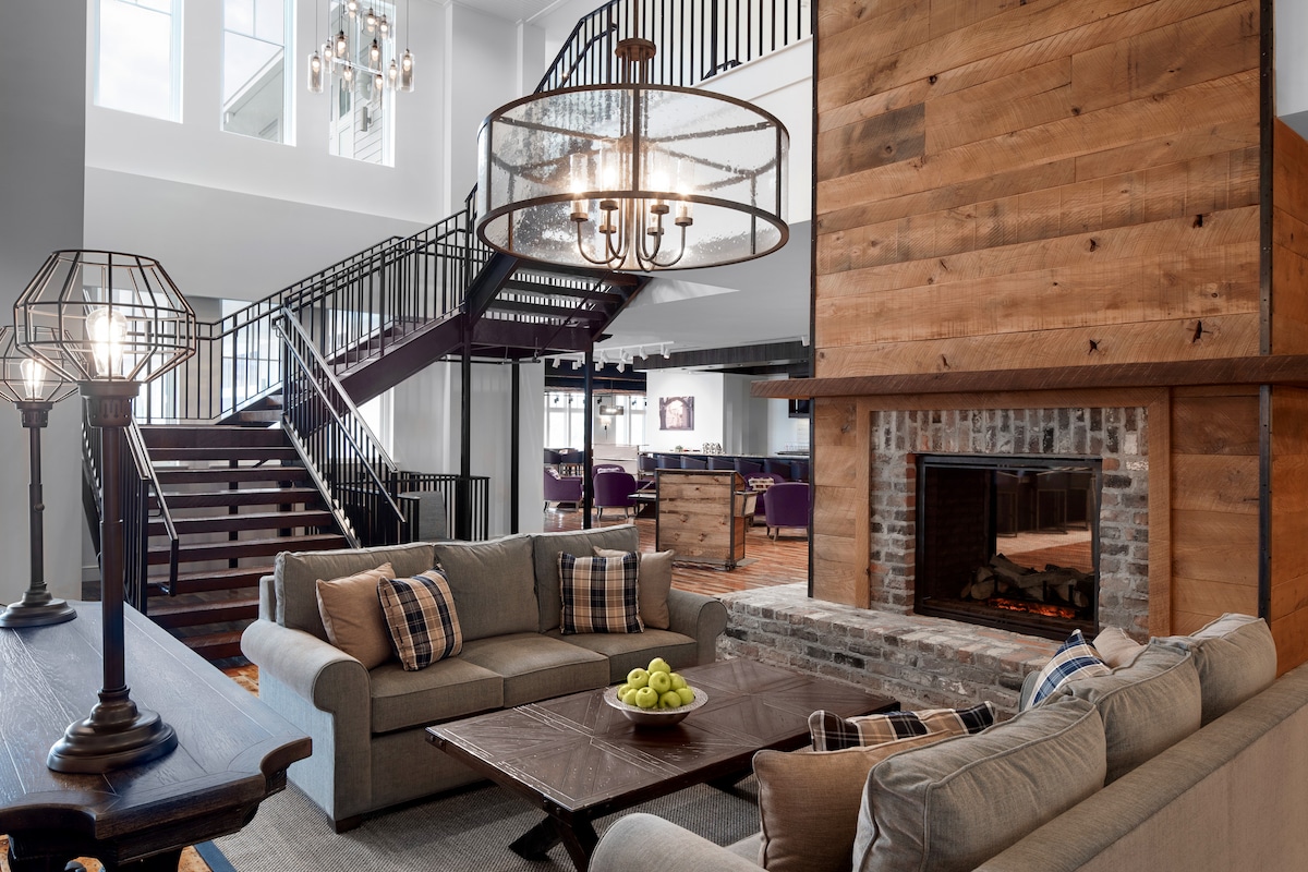 A welcoming lobby area features a warm fireplace framed by rustic brick. Comfortable seating is arranged around a wooden coffee table, with decorative accents including green fruit. An elegant chandelier hangs above, while a spiral staircase leads to the upper level.