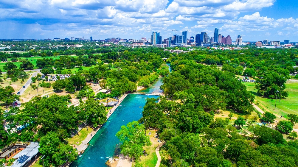 An aerial view captures the lush greenery and winding blue waters of a river, bordered by trees and park areas. In the distance, the skyline of Austin is visible, showcasing a mix of modern buildings and natural landscapes under a partly cloudy sky.