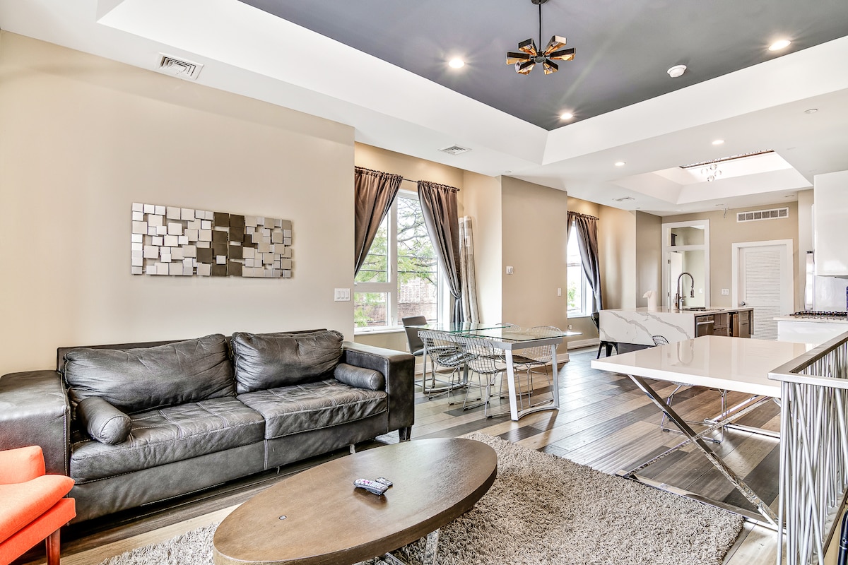 The living area features a dark leather couch paired with an orange accent chair. A modern glass dining table, positioned beside the couch, complements the space. Large windows allow natural light to fill the room, while neutral walls create a calm atmosphere.