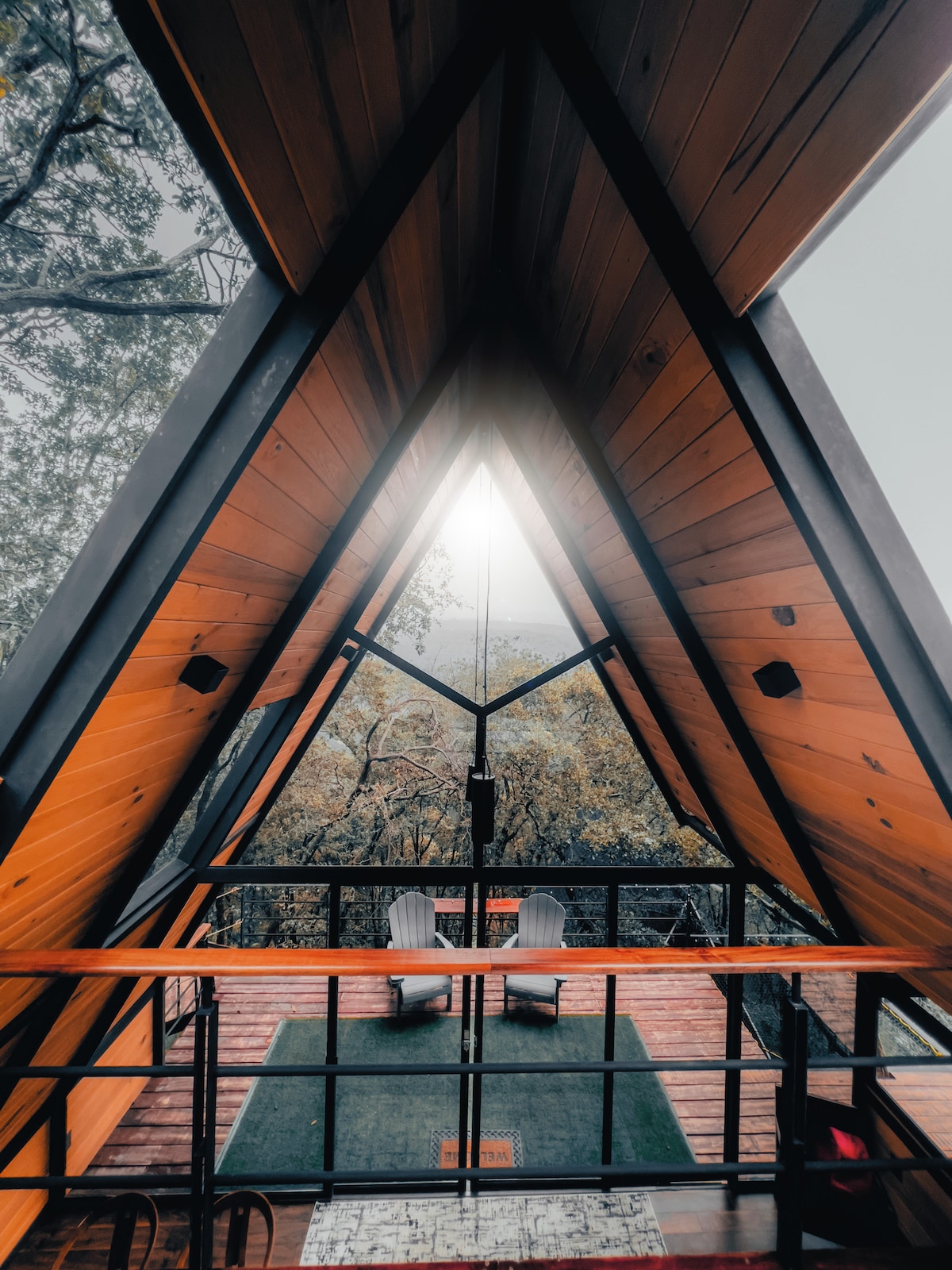 An A-frame style interior is showcased with sloped wooden ceilings, highlighting large windows that frame views of the surrounding forest. Two white chairs are positioned at the edge of a balcony, facing an opening that allows natural light to enter the space.