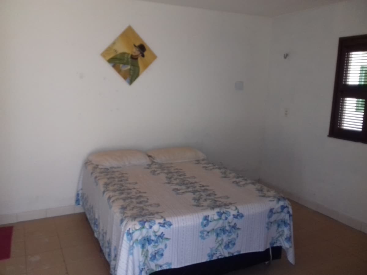 A spacious sleeping area features a double bed dressed in a floral-patterned coverlet. A piece of artwork hangs above the bed, and natural light enters through a window with wooden shutters. The tiled floor and plain walls contribute to a simple, clean environment.