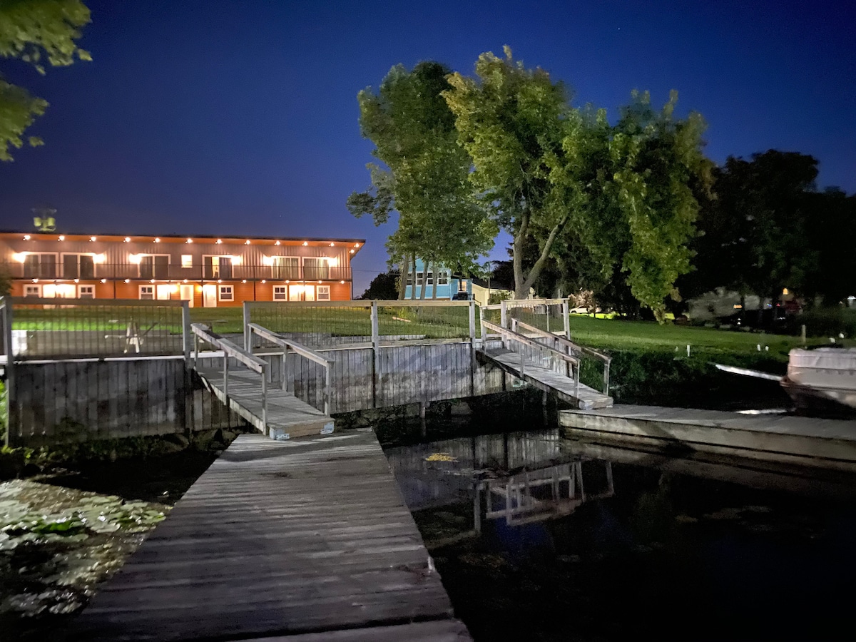 At night, the inviting structure is highlighted by warm lights on the balcony, offering a glimpse of the waterfront lawn and dock area. The boardwalk leads to the water, surrounded by lush greenery and a calm atmosphere, ideal for relaxation.