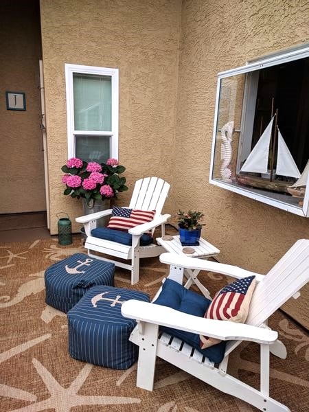 A cozy outdoor seating area features two white Adirondack chairs adorned with decorative pillows. A small table sits between them, accompanied by blue ottomans. A potted plant and vibrant pink flowers are positioned nearby, while a decorative display with a model sailboat is visible in the window.