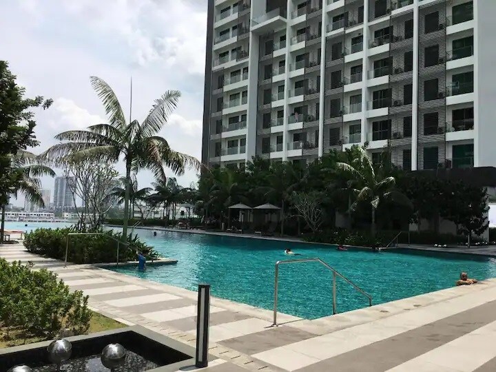 An expansive infinity pool is shown, surrounded by lush palm trees and neatly landscaped gardens. The building's modern architecture is visible in the background, while sun loungers and umbrellas provide shaded areas for relaxation.