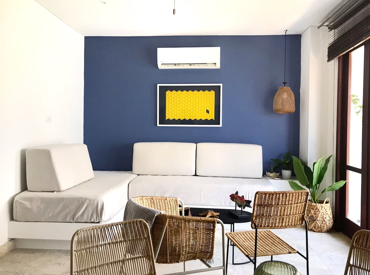 An open living area features a large L-shaped sofa against a deep blue wall, complemented by a vibrant piece of wall art. A selection of woven chairs and a black coffee table create a relaxed seating arrangement. Natural light is welcomed through glass doors, with potted greenery adding a touch of nature.