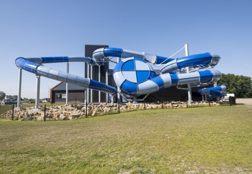 A large water slide structure features multiple twisting and turning blue and white sections. The slides are situated outside against a clear blue sky, with a grassy area visible in the foreground. The surrounding landscape includes a rock formation and parking spaces in the background.