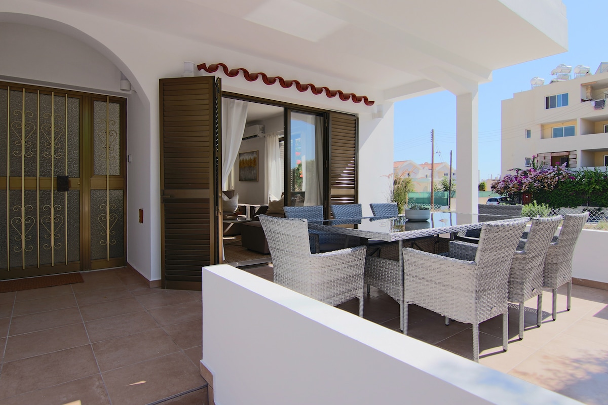 A covered outdoor terrace is presented with a large dining table surrounded by stylish woven chairs. Large sliding doors provide access to an inviting living area, while greenery enhances the view beyond the terrace. The floor features neutral tiles, contributing to a welcoming ambiance.