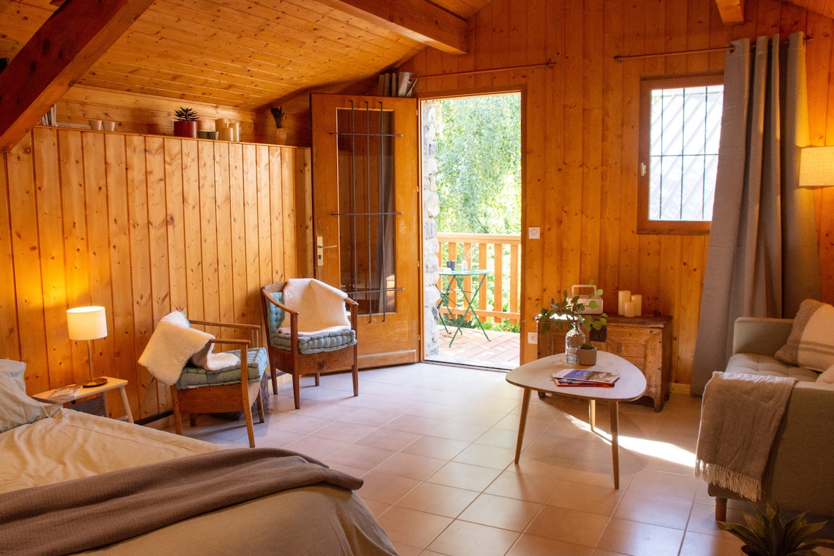 A cozy interior space is showcased, featuring wooden walls and ceiling that enhance the natural ambiance. A spacious bed is positioned alongside a small coffee table and a comfortable sofa. Two armchairs sit near the open door, which leads to a balcony with views of the outdoors.