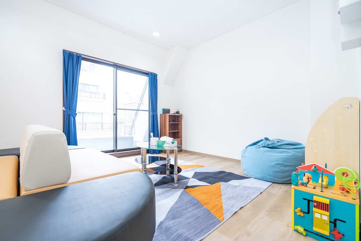 The living space features large windows with blue curtains, allowing ample natural light. A colorful area rug covers the floor, while a compact child-friendly play area is visible. A unique sofa arrangement and bean bag chair provide flexible seating options, making it suitable for families.