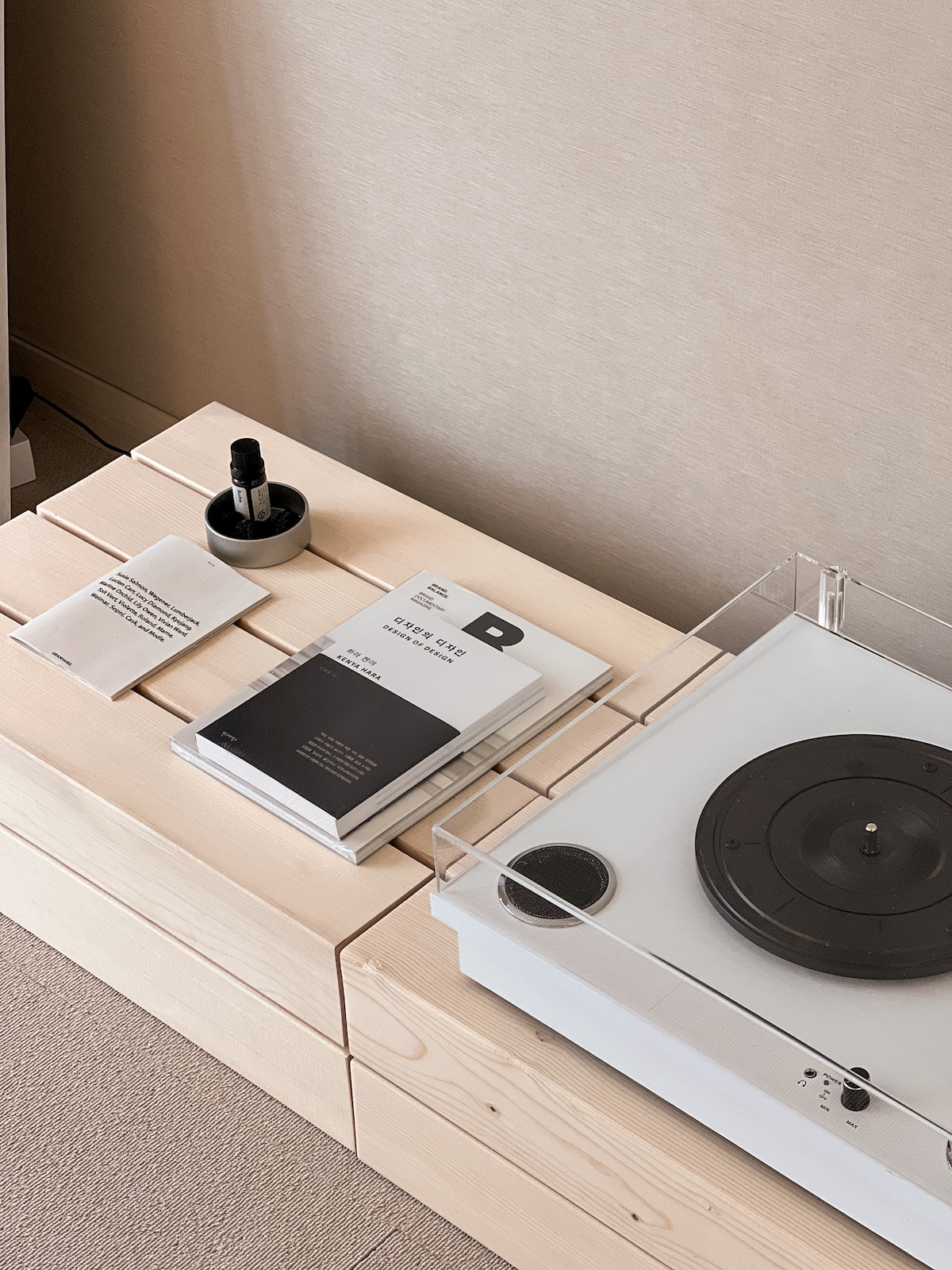 A minimalist wooden table features a turntable and a selection of books placed beside a small decorative item. Soft lighting enhances the simple yet stylish arrangement, inviting relaxation and leisure.