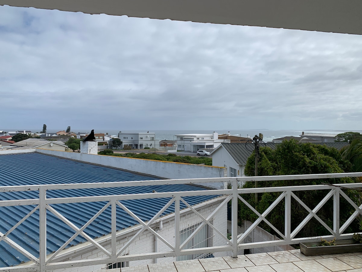A scenic view of the seaside town is captured from the spacious balcony. The image features rooftops and buildings with a glimpse of the ocean in the distance, framed by a white railing and overcast sky.