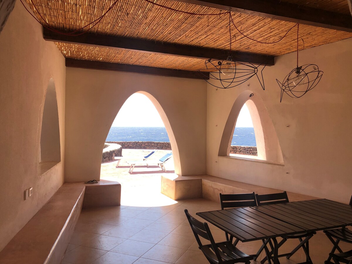 A spacious living area features arched windows that frame a stunning sea view. A large table with black chairs is positioned centrally, while a textured ceiling adds depth. Sun loungers can be seen outside, accessible via a wide entryway.