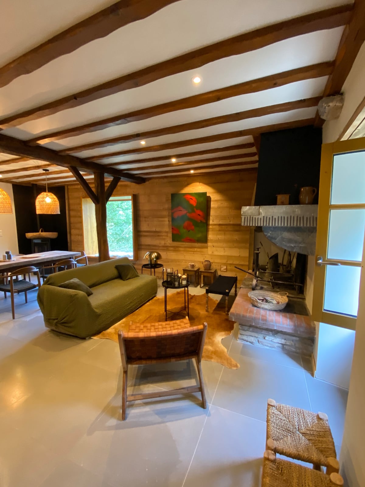 An inviting living area features a green sofa and a rustic fireplace, surrounded by wooden beams overhead. A modern art piece adorns the wall, with natural light filtering through the nearby window. A table and chairs are visible, offering a comfortable space for gatherings.