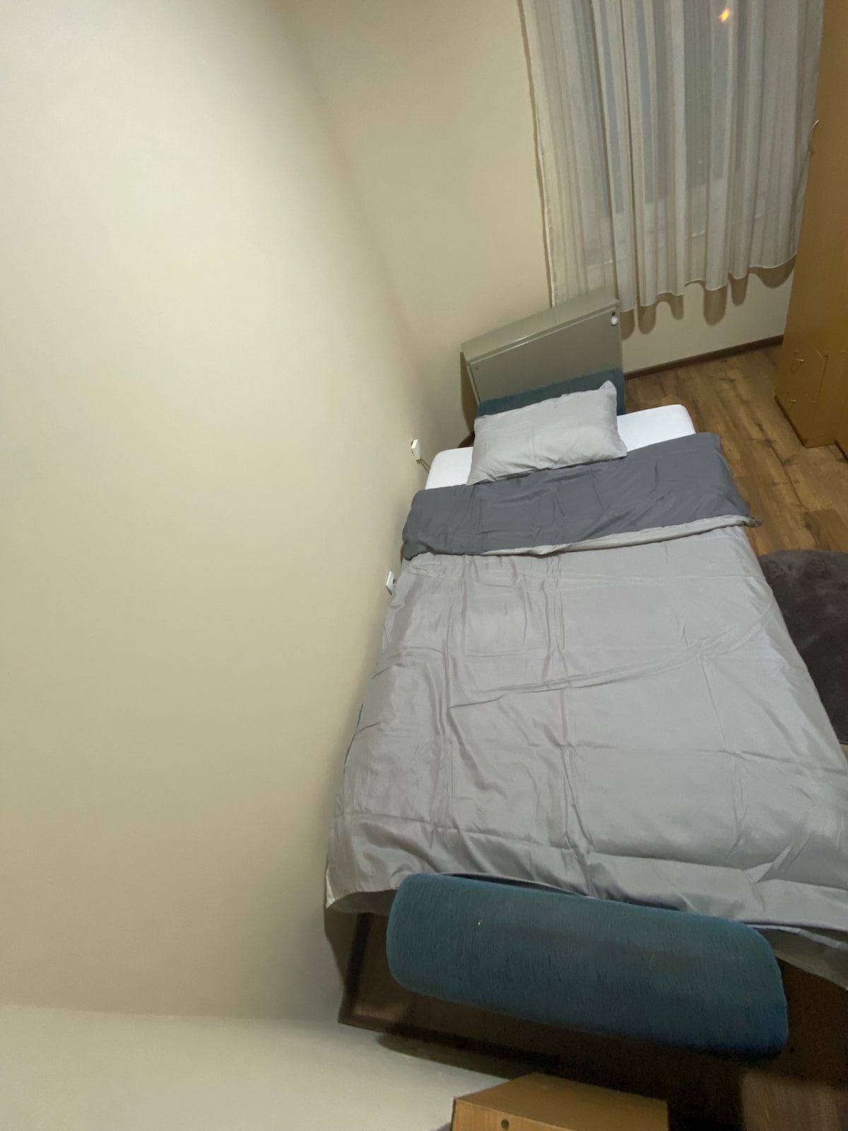 A single bed is positioned against a light-colored wall, covered with a gray blanket and pillow. A wooden floor is visible, and a chair is placed nearby. Natural light enters through the sheer curtains, creating a calm environment.