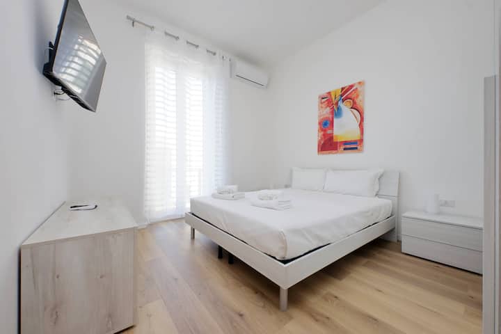 Habitación minimalista y luminosa con cama matrimonial, sábanas blancas impecables, toallas de bienvenida, televisor de pantalla plana en la pared, aire acondicionado, un cuadro abstracto de colores, cortinas transparentes que dejan entrar la luz natural y piso de madera clara.