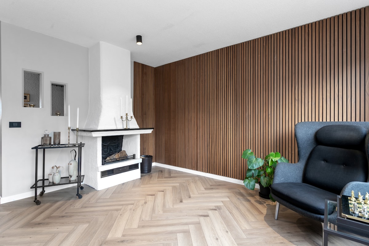 A stylish living room features a modern black fireplace with a wooden accent wall. Natural light filters in through the nearby windows, highlighting the warm herringbone flooring. A comfortable armchair and potted plant complete the inviting atmosphere.