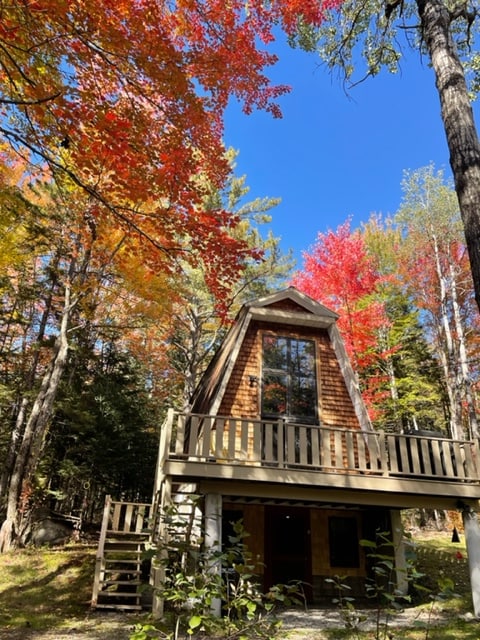 An a-frame cabin is nestled among vibrant autumn foliage. The cabin features a large front balcony and expansive windows, allowing natural light to fill the interior. Tall trees with red and yellow leaves surround the property, creating a picturesque woodland setting.