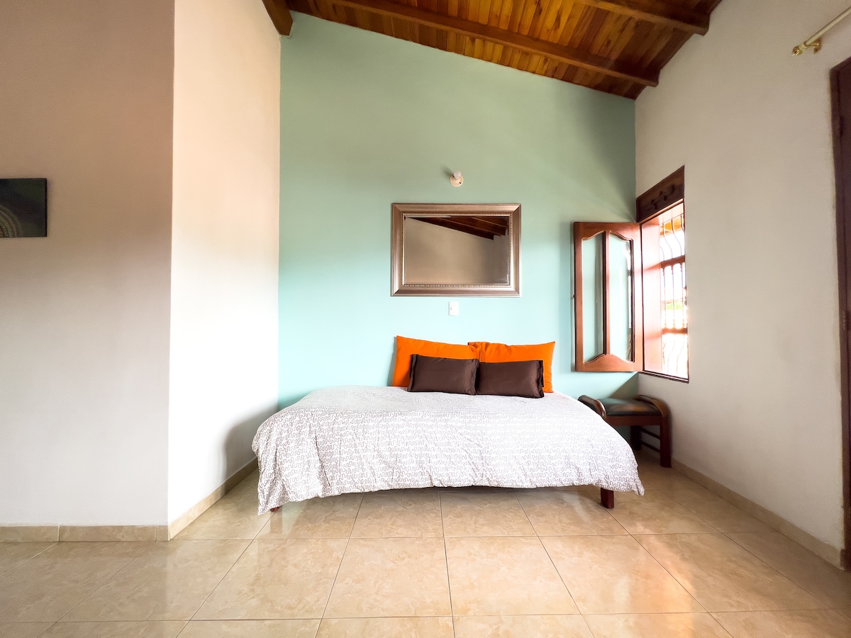 A spacious sleeping area features a large bed dressed with a light coverlet and accent pillows in warm tones. Natural light enters through an open window, and a mirror reflects the calming mint-colored wall. A small chair is positioned next to the bed.