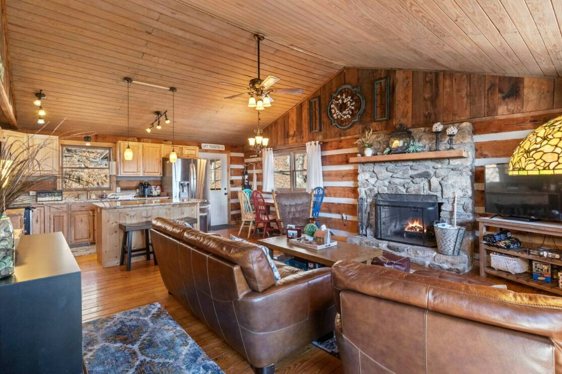A great room is showcased with vaulted ceilings and wooden beams. A stone fireplace is centrally located, surrounded by leather sofas and a patterned rug. The kitchen area is visible in the background, featuring wooden cabinetry and stainless steel appliances.