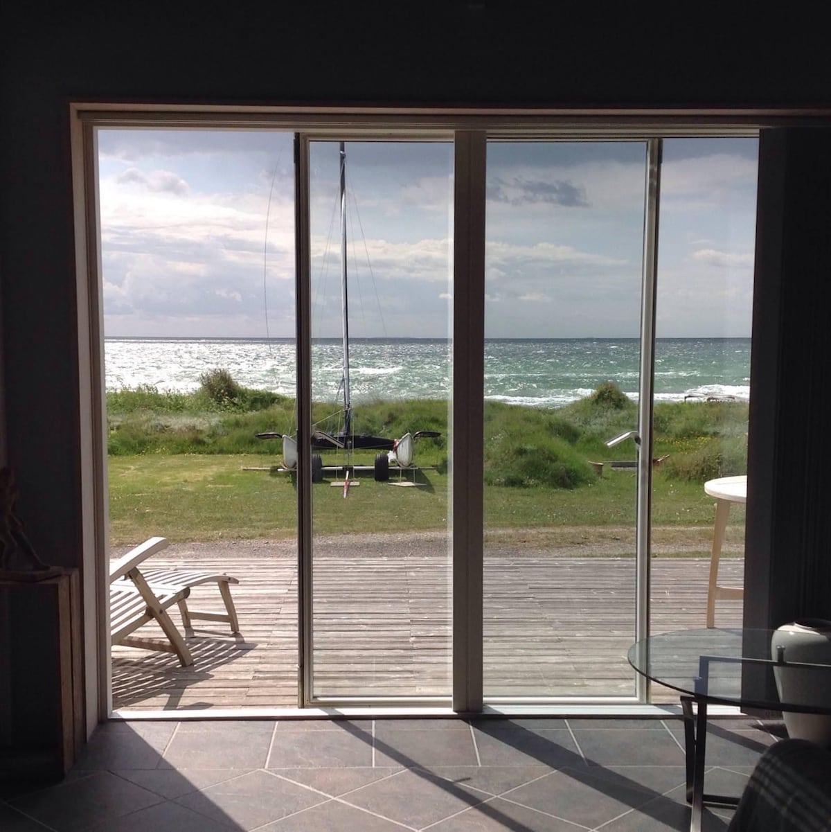 A sliding glass door opens to a view of the sea, where gentle waves meet a grassy shoreline. A sailboat is anchored nearby, and a wooden deck is visible just outside, offering a serene coastal setting.