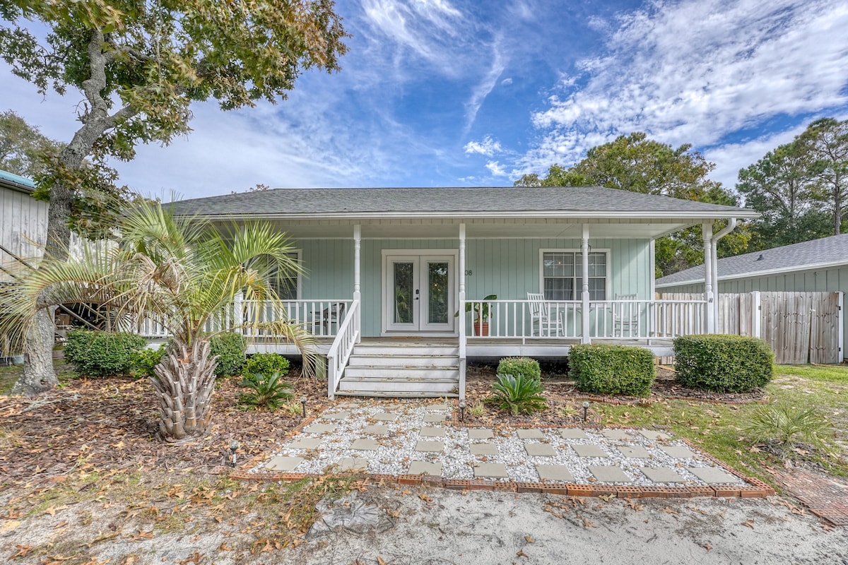 The exterior of a boho-styled beach bungalow is shown, featuring a light blue facade and a welcoming front porch. Two sets of steps lead to the entrance, surrounded by neatly trimmed shrubs and palm trees. A gravel pathway complements the landscaping.