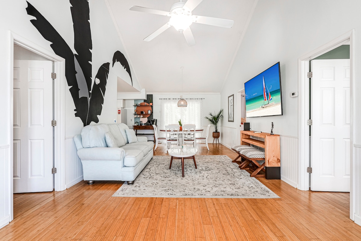 The spacious living area features bamboo flooring, vaulted ceilings, and a hand-painted mural of palm leaves. A light-colored couch sits opposite a large smart TV, with a central coffee table on a textured area rug. A dining table for six is positioned nearby, enhancing the inviting layout.