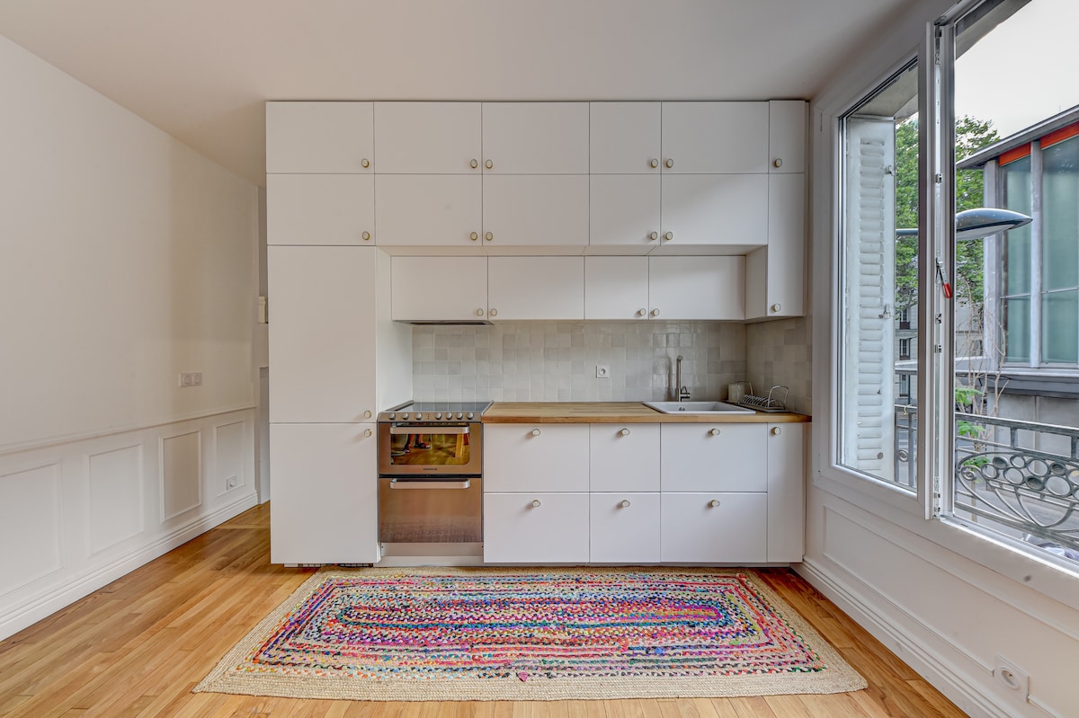 A modern kitchen area showcases sleek white cabinetry and wooden countertops. A built-in oven is integrated into the cabinetry, with a tiled backsplash adding texture. Natural light flows in through a large window, illuminating a colorful woven rug that adds warmth to the hardwood floor.