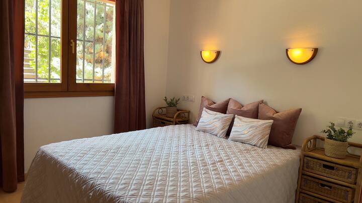 تحتوي غرفة النوم المريحة هذه على سرير مزدوج مع بياضات ناعمة ووسائد وافرة، يضيئها الضوء الطبيعي من نافذة كبيرة. توفر النافذة إطلالة على المنطقة الخارجية والنباتات الخضراء المورقة، مما يخلق أجواء هادئة