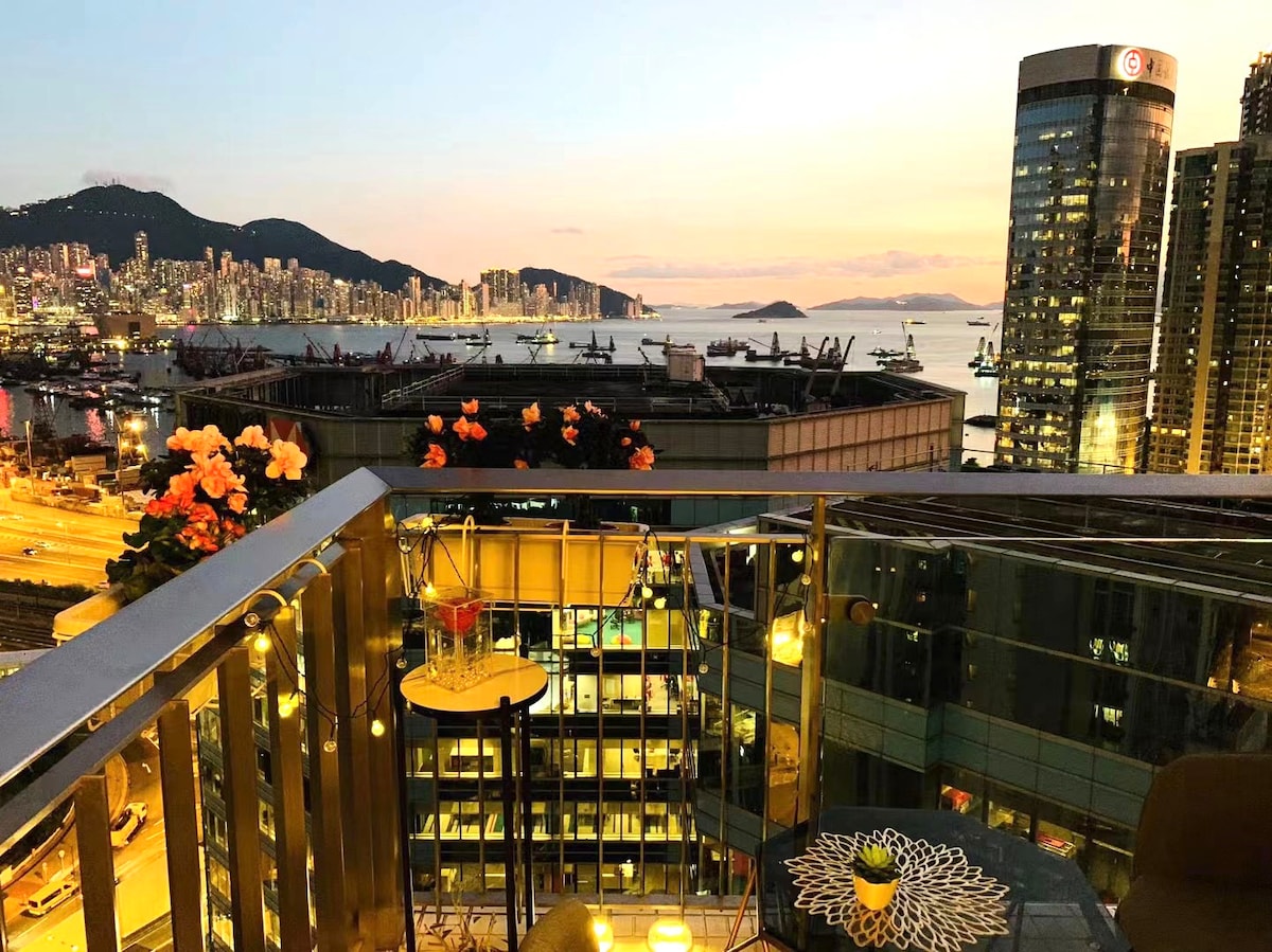 A balcony offers a view of the vibrant city skyline at sunset, with flowers enhancing the ambiance. The scene includes a small table and seating area, surrounded by twinkling lights. The distant mountains and water add depth to the landscape.