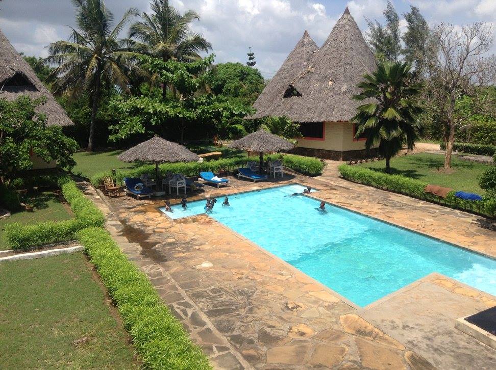 An inviting swimming pool is framed by lush greenery and palm trees. Thatched-roof cabanas provide shaded seating areas around the pool. A well-maintained stone patio surrounds the water, where guests can be seen enjoying the refreshing environment.