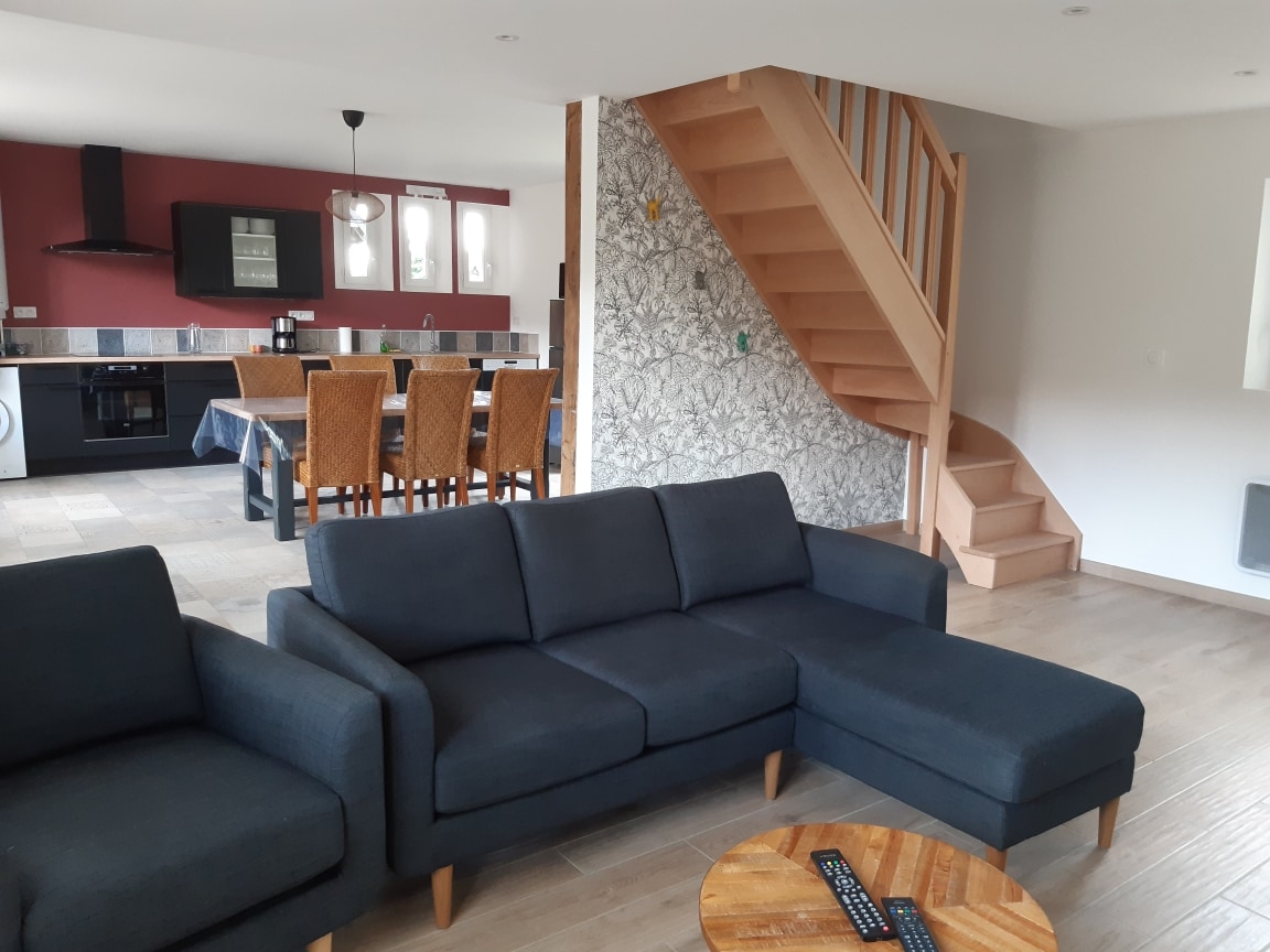 A spacious living area features a dark sectional sofa and a wooden coffee table with a remote control. A rustic dining table is visible in the background, surrounded by wicker chairs. A staircase with a wooden railing leads to the upper level, adding depth to the room.