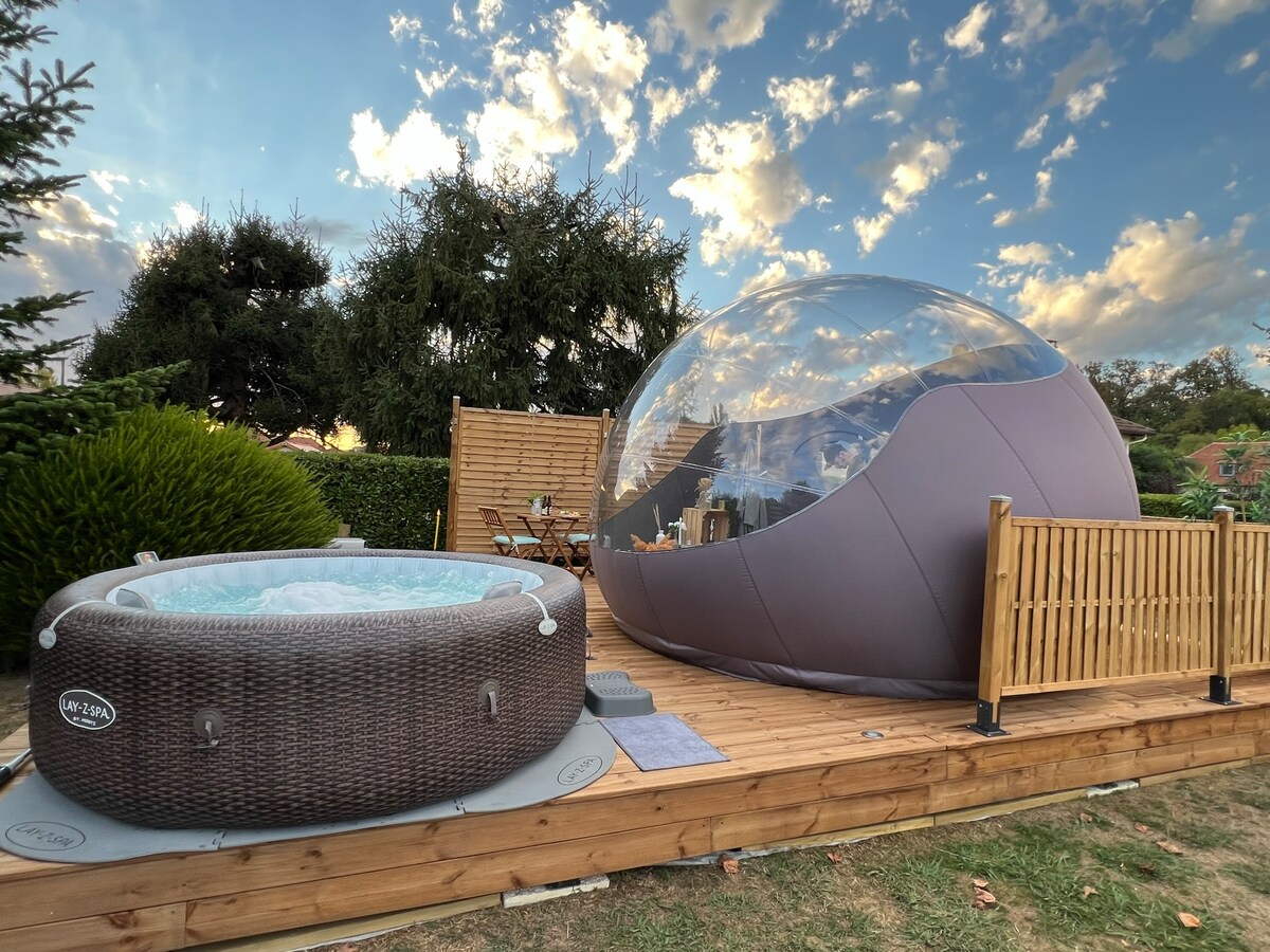 An outdoor area features a bubble accommodation alongside a hot tub. The bubble's transparent design allows for views of the sky, and wooden decking surrounds both the hot tub and bubble. Greenery enhances the tranquil setting, creating a peaceful and immersive environment.