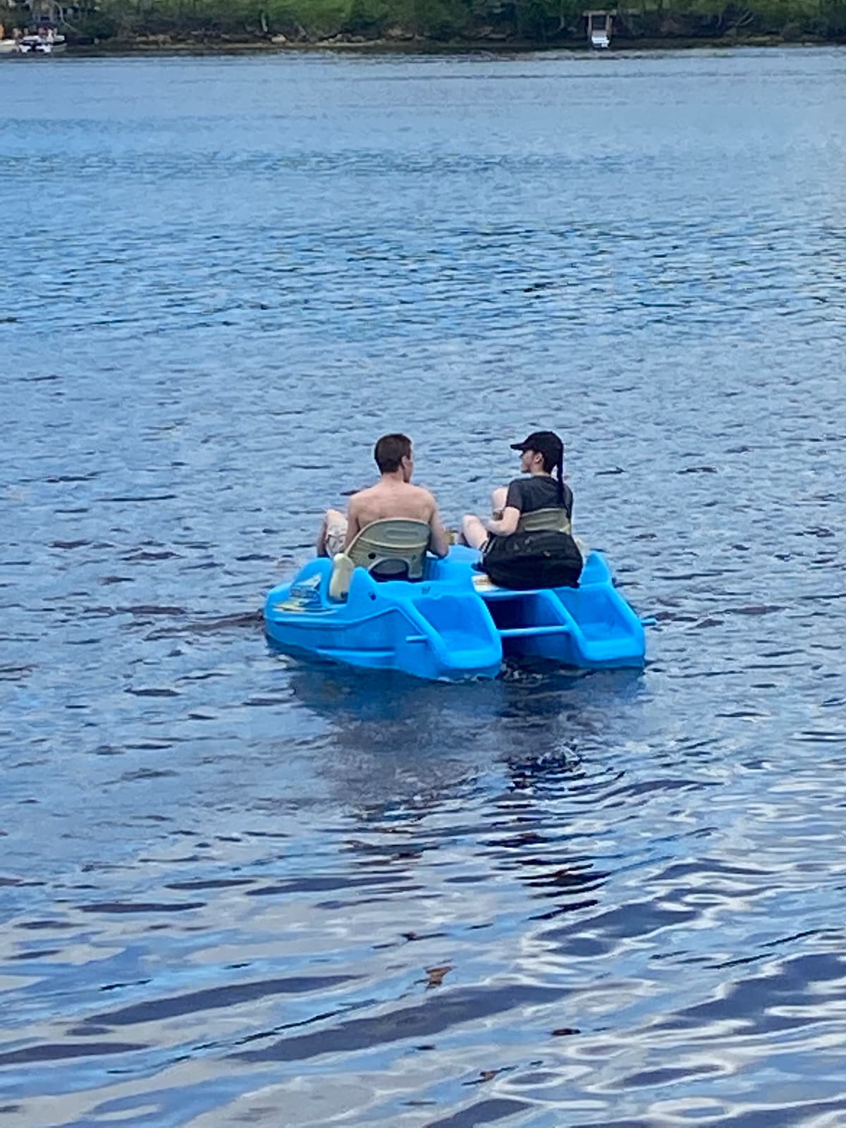 Two individuals are seated on a blue paddle boat, gliding across tranquil waters. The surface of the river reflects surrounding greenery, and gentle ripples can be seen emanating from the boat.